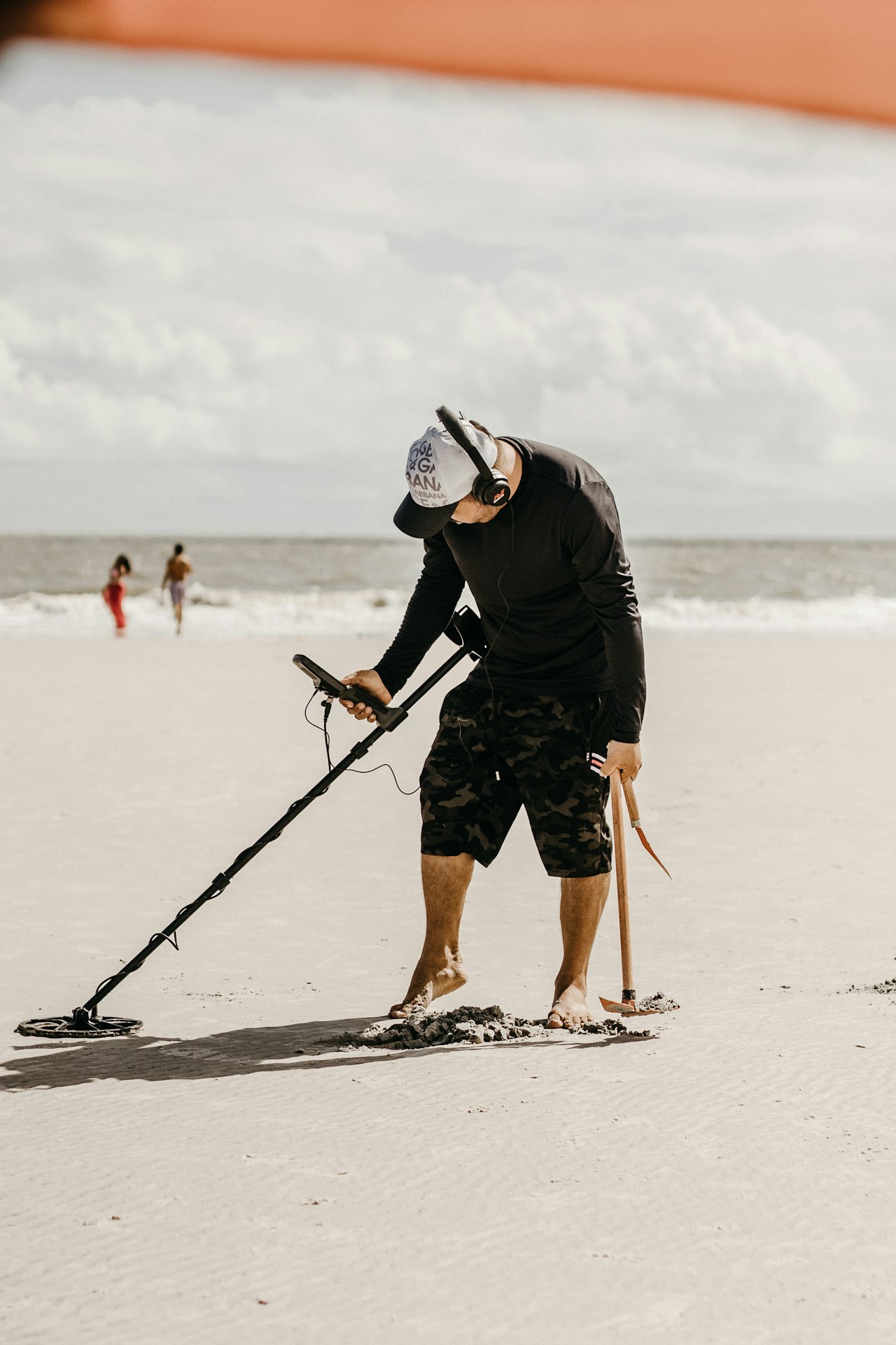 A detectorist swinging a metal detector across wet sand at the water's edge, holding a long-handled sand scoop in his other hand