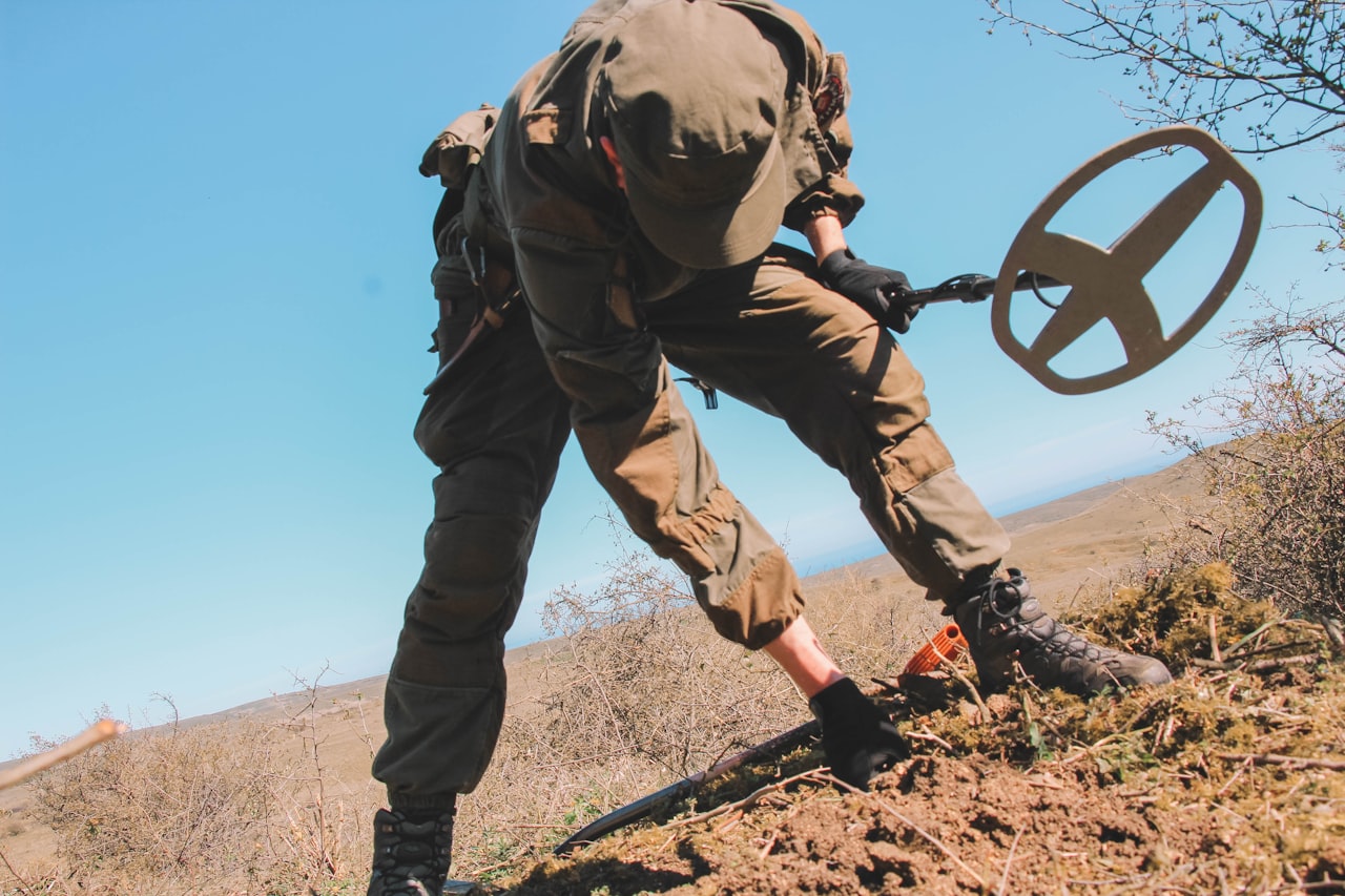A detectorist in cargo pants leaning over a fresh dig with a metal detector and digging tool, hillside in golden light