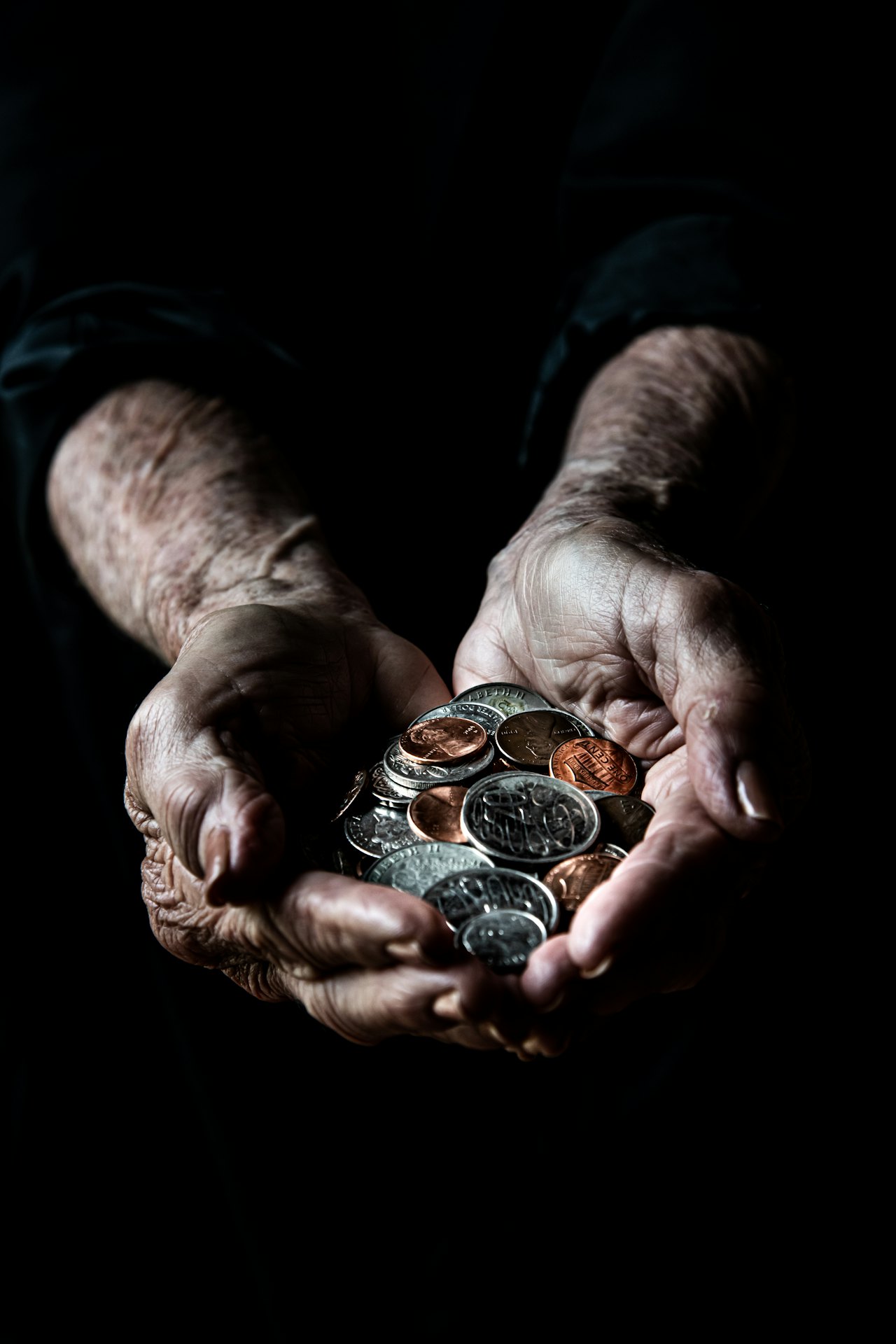 A pair of weathered hands cupping a small pile of silver and copper coins, photographed on a dark background